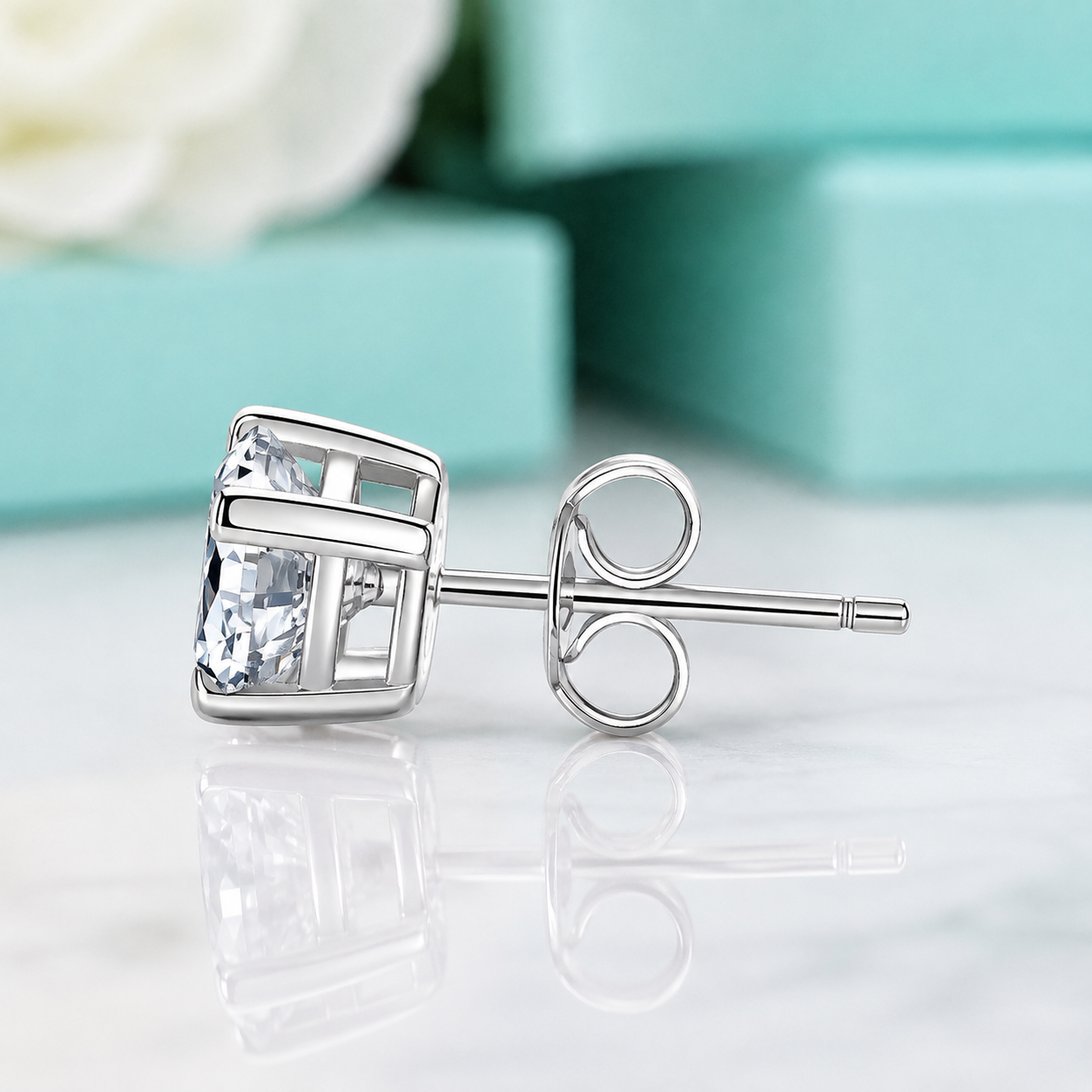 A single silver stud earring with a clear rectangular gemstone, shown on a reflective white surface with blurred turquoise gift boxes in the background.