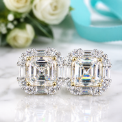 A pair of emerald-cut halo stud earrings in silver with gold accents and baguette side stones, displayed on a white marble surface with white roses and a turquoise ribbon in the background.