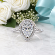 A pear-shaped halo ring with a large clear center stone and pavé-set side stones on a white marble surface, styled with white roses, greenery, and a light blue ribbon in the background.