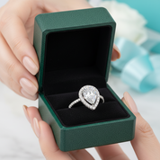 A heart-shaped diamond ring in a silver-toned halo setting, presented inside an open dark green jewelry box held by hands with light pink manicured nails.