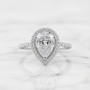 A pear-cut diamond halo ring in white gold or platinum, set on a white marble surface with a soft reflection.