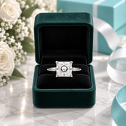 A square princess-cut diamond ring with a blue center stone set in a silver band, displayed inside an open dark green velvet ring box on a marble surface with white roses and a turquoise gift box in the background.