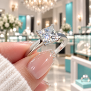 A close-up of a hand holding a silver engagement ring with a clear princess-cut diamond. The background shows a blurred luxury jewelry store interior with teal display cases and warm lighting.