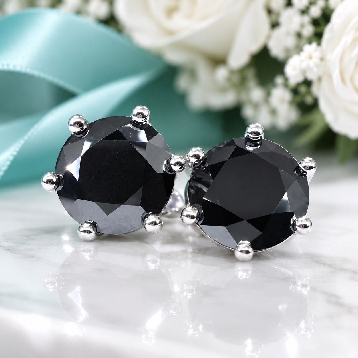 A pair of round stud earrings with faceted black gemstones in silver prong settings, displayed on a white marble surface with a blurred background of white flowers and a teal ribbon.