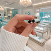 A close-up of a hand with long manicured nails holding a pair of black stud earrings with silver-toned settings, with a blurred jewelry store interior in the background.