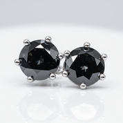 A pair of stud earrings featuring round faceted black gemstones in silver prong settings, displayed on a clean white reflective surface.
