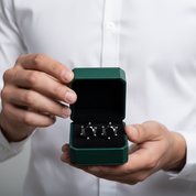 A person in a white shirt holds an open dark green jewelry box displaying a pair of silver-toned earrings with black and white geometric details.