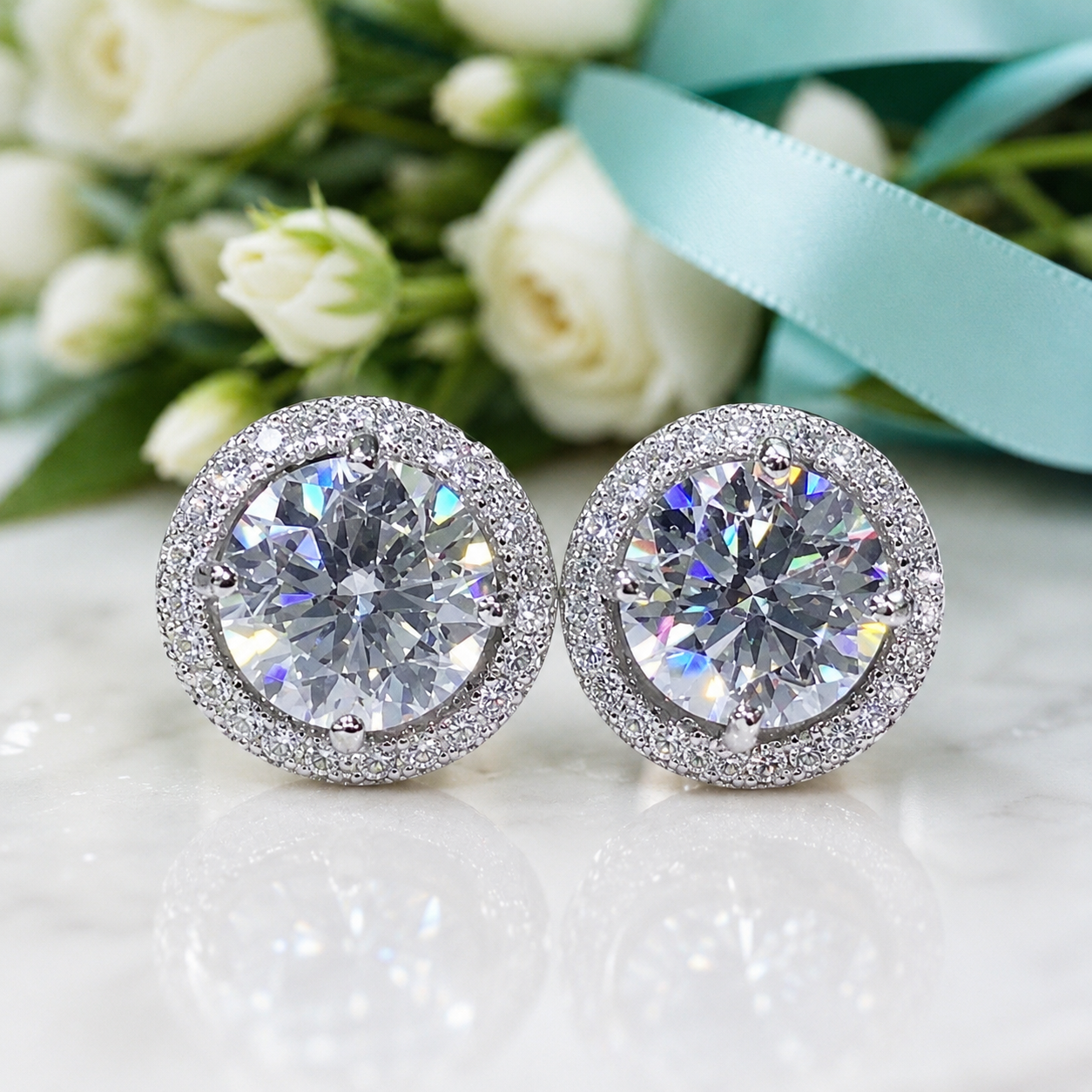 A pair of round stud earrings with large clear center stones surrounded by a halo of small clear stones, displayed on a white reflective surface with white flowers and a light blue ribbon in the background.