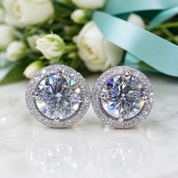 A pair of round stud earrings with large clear center stones surrounded by a halo of small clear stones, displayed on a white reflective surface with white flowers and a light blue ribbon in the background.