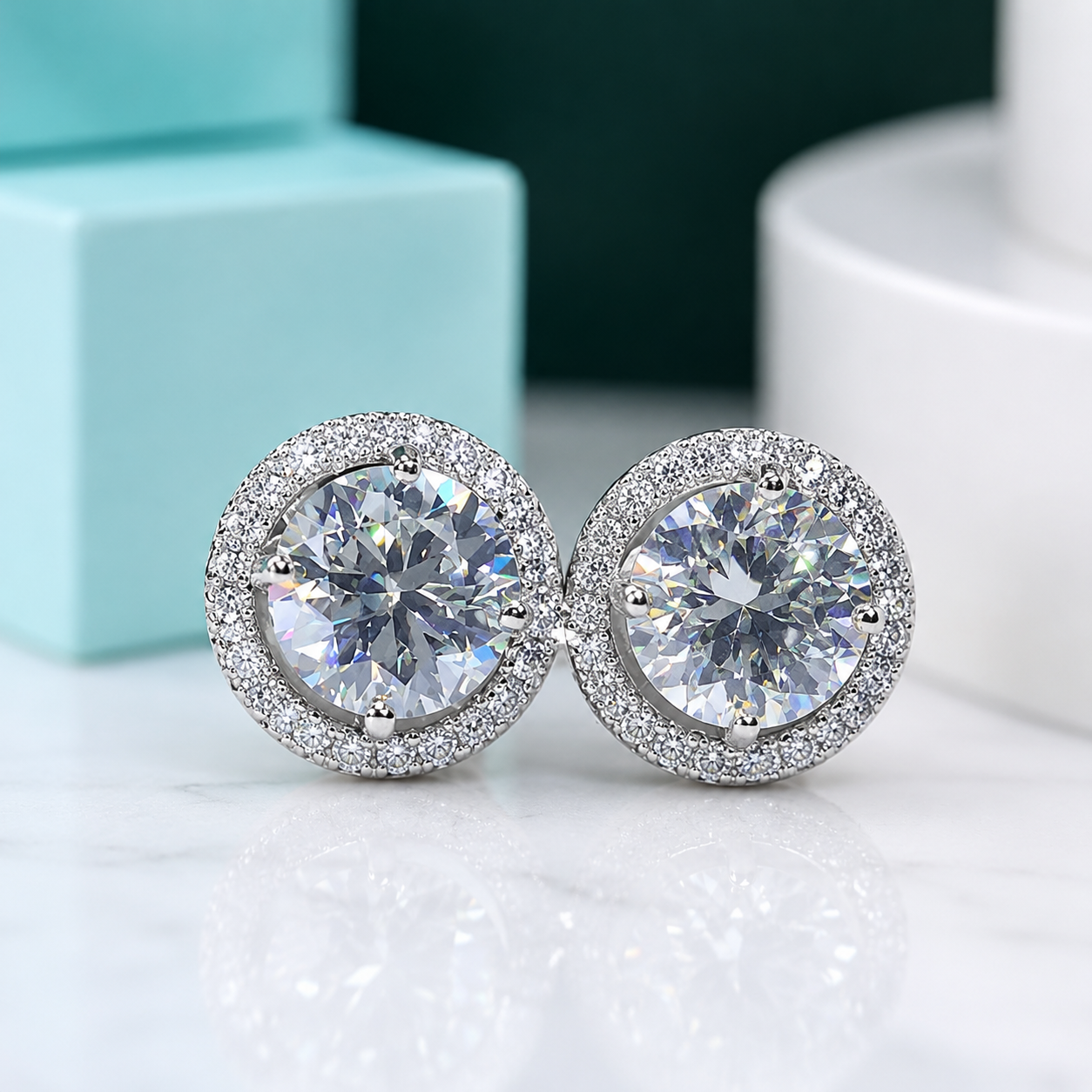 A pair of round stud earrings with large clear center stones surrounded by a halo of smaller clear stones, set in a silver-toned metal, displayed on a light marble surface with a turquoise box and white container in the background.