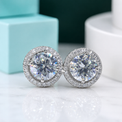 A pair of round stud earrings with large clear center stones surrounded by a halo of smaller clear stones, set in a silver-toned metal, displayed on a light marble surface with a turquoise box and white container in the background.