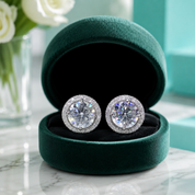 A pair of round halo stud earrings with clear center stones and pavé settings, displayed in an open dark green velvet jewelry box on a marble surface.