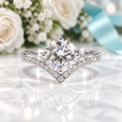 A radiant cut diamond engagement ring with a V-shaped pavé band, set on a white marble surface with blurred white roses and a light blue ribbon in the background.