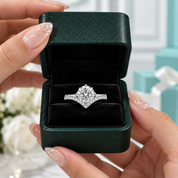 A silver engagement ring with a round-cut center stone and pavé-set side stones is displayed inside an open dark green textured jewelry box, held by hands with manicured nails.