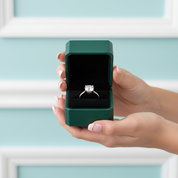 A silver engagement ring with a round-cut clear gemstone is displayed inside an open dark green jewelry box held by two hands against a light blue paneled background.