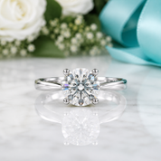 A classic solitaire engagement ring with a round brilliant-cut diamond set in a polished silver-colored band, resting on a white marble surface with a soft reflection. The background is decorated with white roses, baby's breath, and a light blue ribbon.