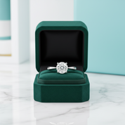 A round-cut diamond engagement ring in a silver band, displayed inside an open dark green velvet jewelry box on a white marble surface.