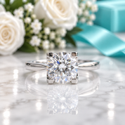 A classic solitaire engagement ring with a round brilliant-cut diamond set in a silver-colored band, resting on a polished white marble surface with a soft reflection. The background is a soft-focus floral arrangement with white roses, baby's breath, and a turquoise ribbon.