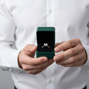 A person in a white dress shirt holds an open dark green ring box displaying a silver ring with a clear center stone.