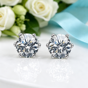 A pair of round-cut silver stud earrings with clear faceted gemstones, displayed on a white reflective surface with white roses and a light blue ribbon in the background.