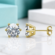A pair of yellow gold stud earrings with round brilliant-cut clear gemstones, shown on a reflective white surface with a blurred turquoise and white background.