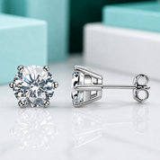 A pair of silver stud earrings, one showing the front with a round brilliant-cut clear gemstone in a four-prong setting and the other showing the side profile with the post and butterfly back, displayed on a reflective white surface with blurred turquoise and white boxes in the background.