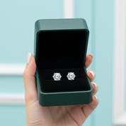 A hand holds an open dark green jewelry box with a black velvet interior, displaying a pair of round-cut silver stud earrings against a soft light blue background.