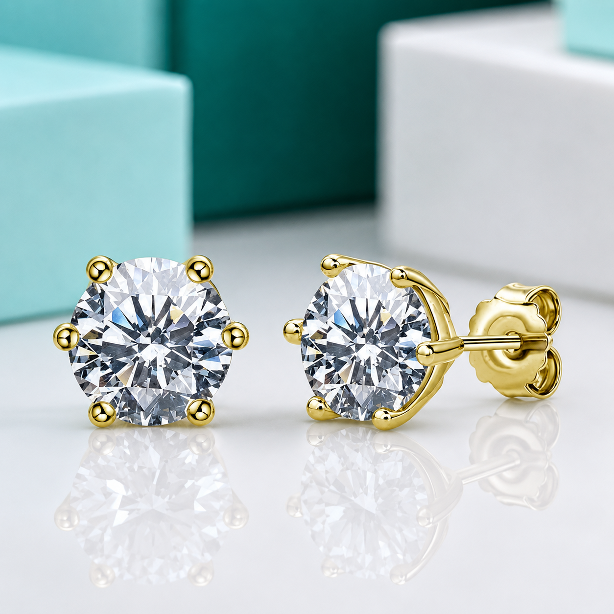 A pair of yellow gold stud earrings with round brilliant-cut clear gemstones in a prong setting, shown on a reflective white surface with blurred turquoise and white boxes in the background.