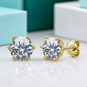 A pair of yellow gold stud earrings with round brilliant-cut clear gemstones in a prong setting, shown on a reflective white surface with blurred turquoise and white boxes in the background.