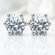 A pair of round-cut diamond stud earrings in a six-prong setting with silver-toned metal, displayed on a reflective white surface against a soft white background.