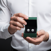 A person in a white dress shirt holds an open dark green jewelry box displaying a pair of round silver-toned stud earrings with clear gemstones.