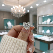 A heart-shaped diamond engagement ring with a silver-colored band held by a hand in a jewelry store.