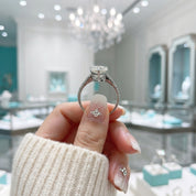 A hand with glittery manicured nails holds a silver-toned diamond engagement ring in front of a blurred luxury jewelry store background with Tiffany-style boxes and a chandelier.
