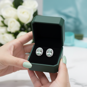 A pair of round stud earrings with large clear center stones and a halo of smaller clear stones, displayed inside an open dark green jewelry box held by hands with mint green nail polish. White roses and a soft blue background are visible.