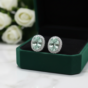 A pair of oval stud earrings with large pale green center stones surrounded by small clear stones, displayed on a dark green jewelry box with white roses blurred in the background.
