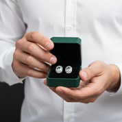 A person in a white button-up shirt holds an open dark green jewelry box displaying a pair of round silver-toned stud earrings with clear stones.