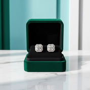 A pair of square-cut diamond stud earrings displayed in an open dark green velvet jewelry box on a white marble surface.