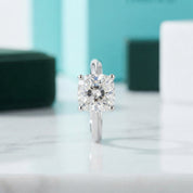 A square-cut solitaire diamond ring in a silver-toned setting, standing upright on a light marble surface with a blurred turquoise and white background.