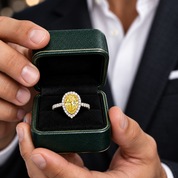 A close-up lifestyle shot of a man holding an open dark green ring box. Inside the box is a yellow diamond engagement ring with a halo setting.