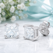 A pair of princess-cut clear gemstone stud earrings in a silver-tone setting, shown on a reflective white surface with soft white flowers and a light blue satin bow in the background.