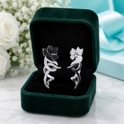 A pair of silver-toned floral earrings with pavé-set clear stones, displayed inside an open dark green velvet jewelry box on a light marble surface with white flowers and a light blue gift box in the background.