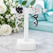 A pair of silver and black winged fairy earrings displayed on a white T-stand. The earrings feature clear pavé-set stones that create a white/silver appearance, with one earring accented by black stones.