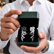 A person in a white shirt holds an open dark green jewelry box displaying a pair of silver floral ring stands with sparkling clear stones.