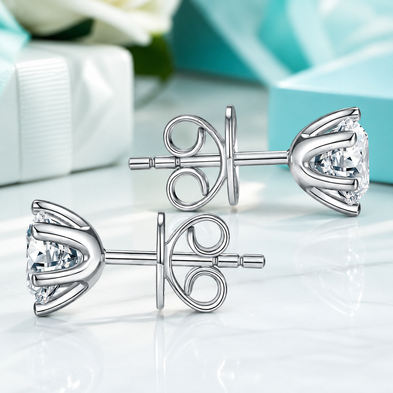 A pair of silver-toned stud earrings with large clear gemstones in a floral or tulip-like setting, shown on a glossy white surface with soft turquoise and white gift-box props in the background.