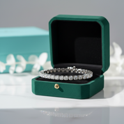 A diamond tennis bracelet displayed inside an open dark green jewelry box on a white surface, with a blurred turquoise box and white floral decor in the background.
