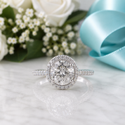 A silver halo engagement ring with a round center stone and pavé band, displayed on a white marble surface with white roses, baby's breath, and a light blue ribbon in the background.