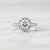 A round-cut halo engagement ring with a brilliant center stone and a surrounding halo of small pavé-set diamonds, set on a silver-toned band with additional diamonds, resting on a white marble surface with a soft reflection.