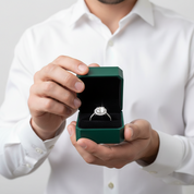 A silver diamond engagement ring displayed inside an open dark green presentation box, held by a person wearing a white dress shirt.