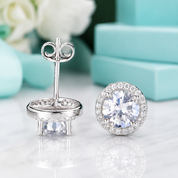 A pair of round halo stud earrings in silver-tone metal with clear gemstones, shown on a white marble surface with a turquoise gift box and blurred flowers in the background. One earring is shown from the front and the other from the back.