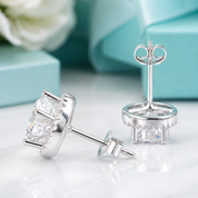 A pair of silver stud earrings with three clear rectangular-cut stones in a halo setting, shown on a white marble surface with a blurred turquoise gift box and white flower in the background.
